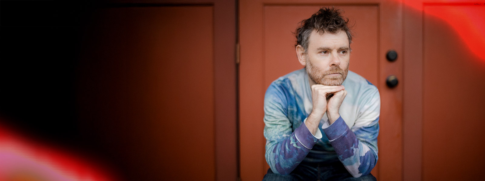 Gabriel Kahane sitting in front of a red wall with his hands on his chin and knees