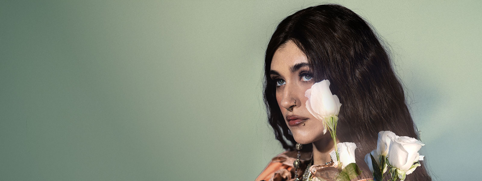 Sierra Ferrell looking up into the distance in front of a dreamy light green background with white roses covering part of her face and hair