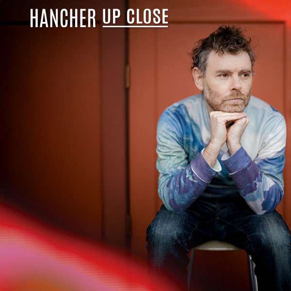 Gabriel Kahane sitting in front of a red wall with his hands on his chin and knees