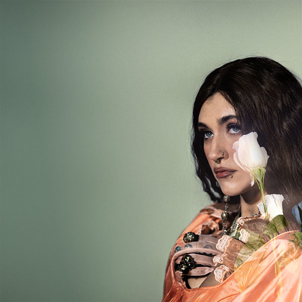 Sierra Ferrell looking up into the distance in front of a dreamy light green background with white roses covering part of her face and hair