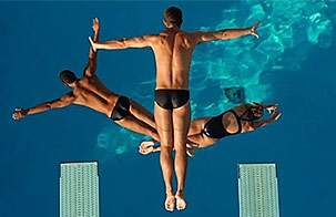 Three high divers in swimsuits from Dana Kunze’s Watershow Productions are photographed mid-dive as they jump from diving boards into the blue water of swimming pool far below them.