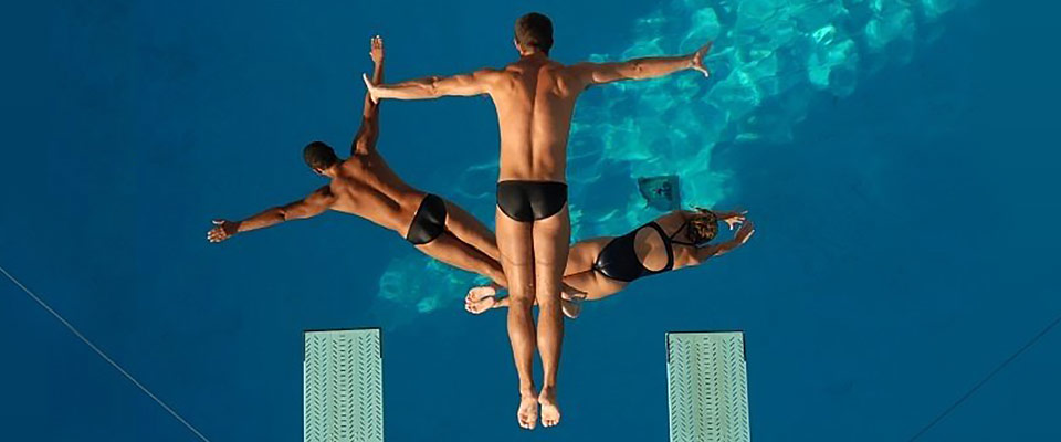 Three high divers in swimsuits from Dana Kunze’s Watershow Productions are photographed mid-dive as they jump from diving boards into the blue water of swimming pool far below them.