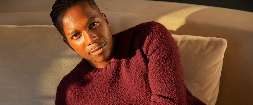 Leslie Odom Jr. wearing a burgundy top, sitting on a cream-colored couch