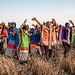 Many individuals singing and dancing in colorful outfits in the tall grass