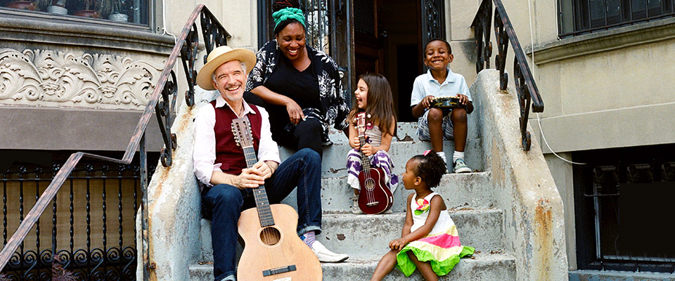 Dan + Claudia Zanes sit on the stoop of a brownstone home, sharing a laugh with three children