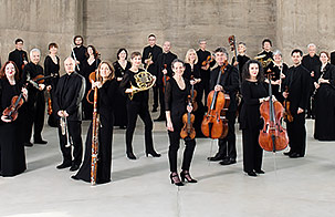The musicians of Academy of St Martin in the Fields stand against a concrete background with their instruments, smiling at the camera