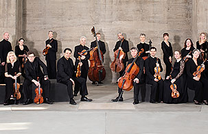 The musicians of Academy of St Martin in the Fields pose with their string intstruments in a modern, concrete space
