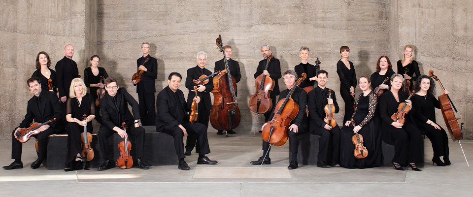 The musicians of Academy of St Martin in the Field pose with their string intstruments in a modern, concrete space