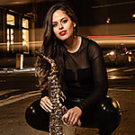 Alexa Tarantino, dressed in a black and holding her saxophone, crouches on the pavement of a city intersection at nighttime