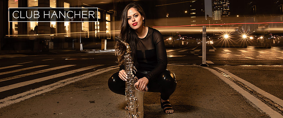 Alexa Tarantino, dressed in a black and holding her saxophone, crouches on the pavement of a city intersection at nighttime