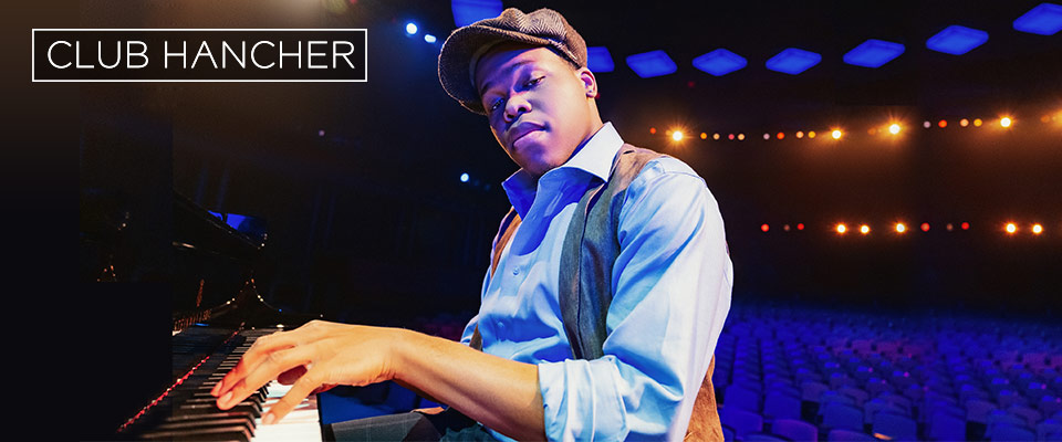 Isaiah J. Thompson, wearing a newsboy cap and a vest, looks at the camera confidently while playing piano