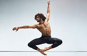 Alvin Ailey male dancer in leaping pose with toes pointed together and knees out with left arm above his head and right arm following his eyes to the right