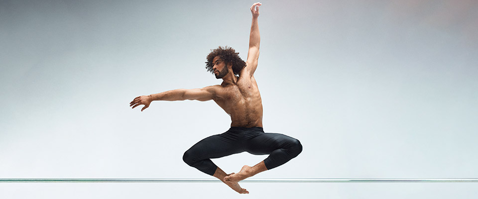 Alvin Ailey male dancer in leaping pose with toes pointed together and knees out with left arm above his head and right arm following his eyes to the right