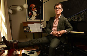 Ben Folds sitting at at a piano backwards. Wearing a dark suit with a pink button up underneath and glasses
