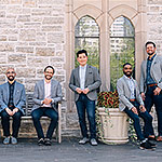 The members of Cantus pose against a light-colored brick wall in jeans and sport coats