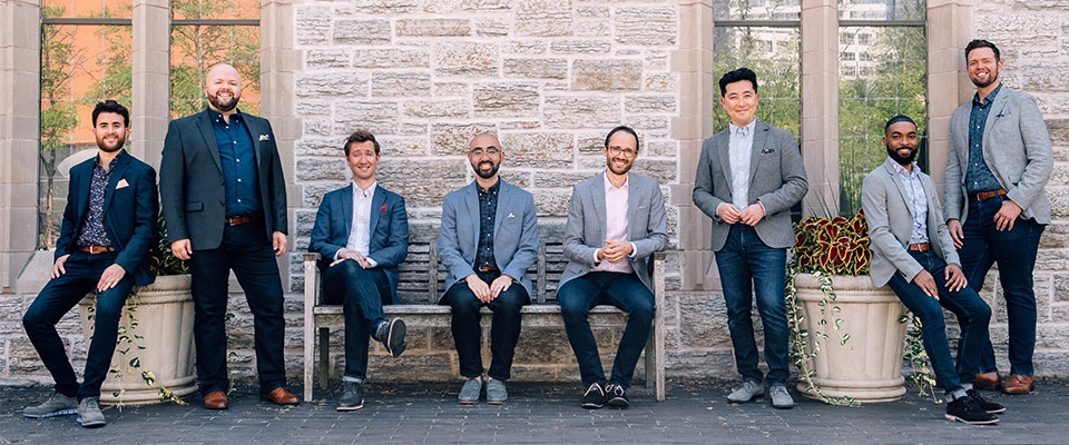 The Cantus boys pose against a light-colored brick wall in jeans and sport coats
