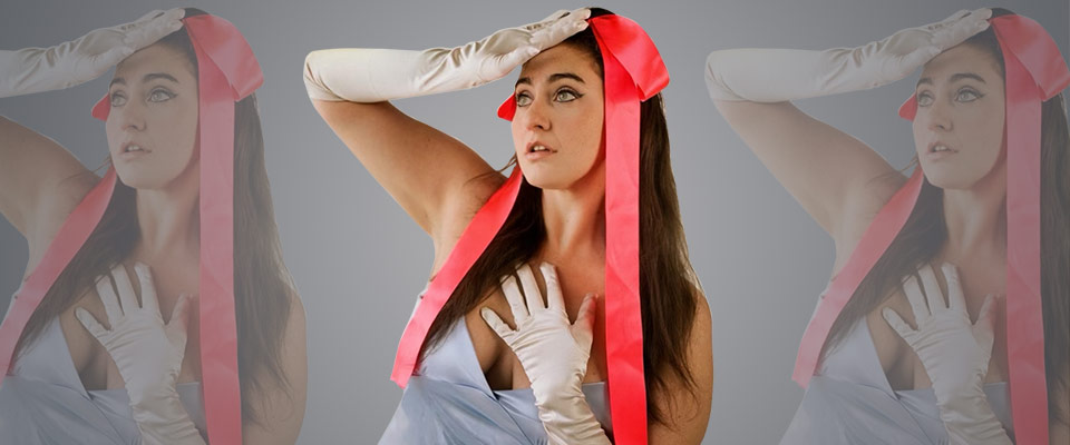Cat Cohen in a silver dress with a large pink bow on her head. She is touching the top of the bow with her right hand and left hand is placed against her chest. She is looking up and to the left of the image. Shadowed images on left and right side