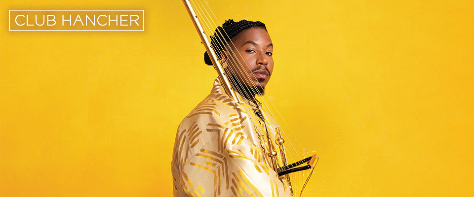 Chief Xian aTunde Adjuah wearing gold outfit against a gold background. He is standing facing right, but looking straight on at the camera. Club Hancher text treatment in upper left corner