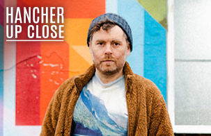 Gabriel Kahane: Magnificent Bird & Book of Travelers Gabriel Kahane standing in front of a colorful wall wearing a sweater and a blue knit hat