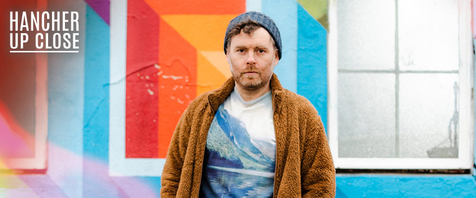 Gabriel Kahane: Magnificent Bird & Book of Travelers Gabriel Kahane standing in front of a colorful wall wearing a sweater and a blue knit hat