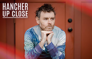 Gabriel Kahane: Magnificent Bird & Book of Travelers Gabriel Kahane resting his hands on his head against a red background