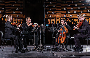 JACK Quartet sitting in half circle in all black clothing playing their instruments on stage