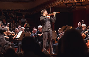 Joshua Bell and Academy of St. Martin in the Fields Joshua Bell playing while standing in the middle of an ensemble sitting down and playing string instruments