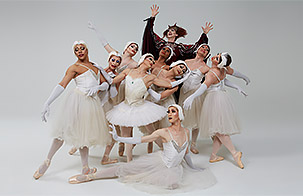 Les Ballets Trockadero de Monte Carlo posing with a white background. All members wearing white costumes. One performer in back wearing dark costume. 