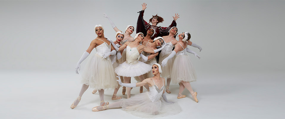 Les Ballets Trockadero de Monte Carlo posing with a white background. All members wearing white costumes. One performer in back wearing dark costume. 