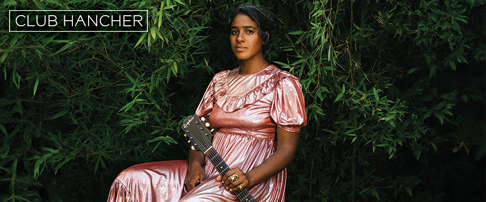 Leyla McCalla sitting in a pink dress against greenery holding her guitar to her left side. Club Hancher text treatment in upper left corner