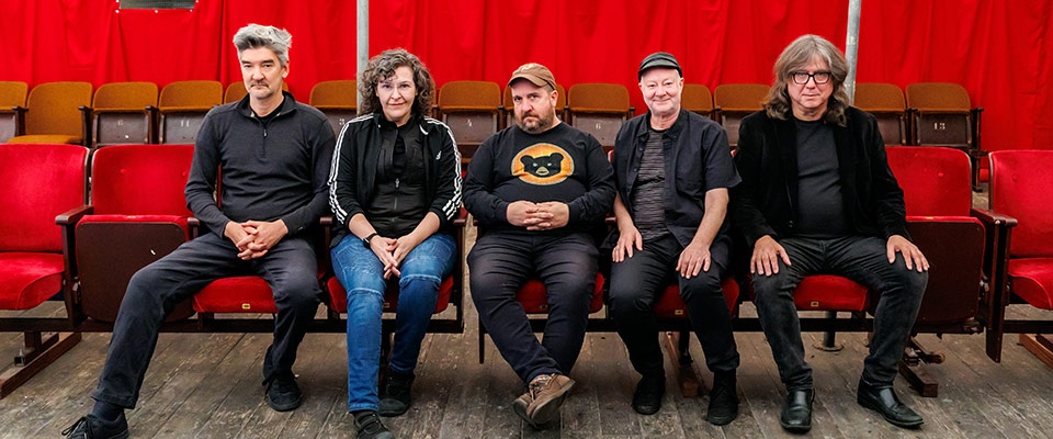 Magnetic Fields band members sitting in a line in red auditorium seats. All members are wearing black shirts and looking at the camera