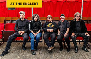 Magnetic Fields band members sitting in a line in red auditorium seats. All members are wearing black shirts and looking at the camera