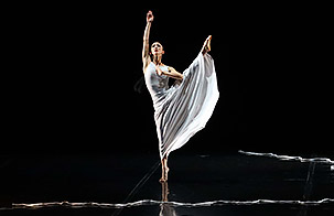 Martha Graham Dancer center stage in white costume with left leg extended towards ceiling against the contrast of black stage