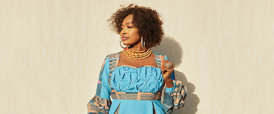 Oumou Sangaré center focus looking to the left against a tan background wearing light blue, yellow, and red colors. 