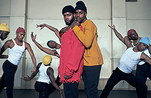 serpentwithfeet dancers in white tank tops and colored stocking hats in movement in background with two members standing in front and center looking into the camera, one embracing the other from behind. 
