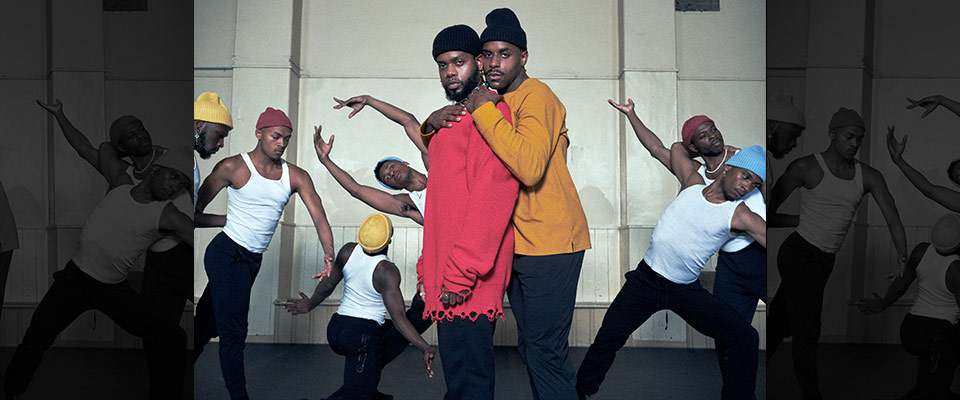 serpentwithfeet dancers in white tank tops and colored stocking hats in movement in background with two members standing in front and center looking into the camera, one embracing the other from behind. 