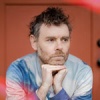 Gabriel Kahane resting his hands on his head against a red background