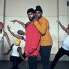 serpentwithfeet dancers in white tank tops and colored stocking hats in movement in background with two members standing in front and center looking into the camera, one embracing the other from behind. 