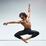 Alvin Ailey male dancer in leaping pose with toes pointed together and knees out with left arm above his head and right arm following his eyes to the right