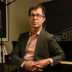 Ben Folds sitting at at a piano backwards. Wearing a dark suit with a pink button up underneath and glasses