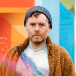 Gabriel Kahane: Magnificent Bird & Book of Travelers Gabriel Kahane standing in front of a colorful wall wearing a sweater and a blue knit hat