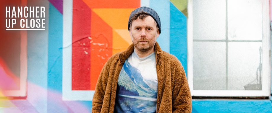 Gabriel Kahane: Magnificent Bird & Book of Travelers Gabriel Kahane standing in front of a colorful wall wearing a sweater and a blue knit hat