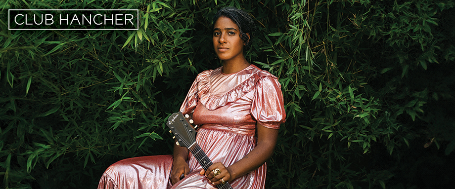 Leyla McCalla sitting in a pink dress against greenery holding her guitar to her left side. Club Hancher text treatment in upper left corner